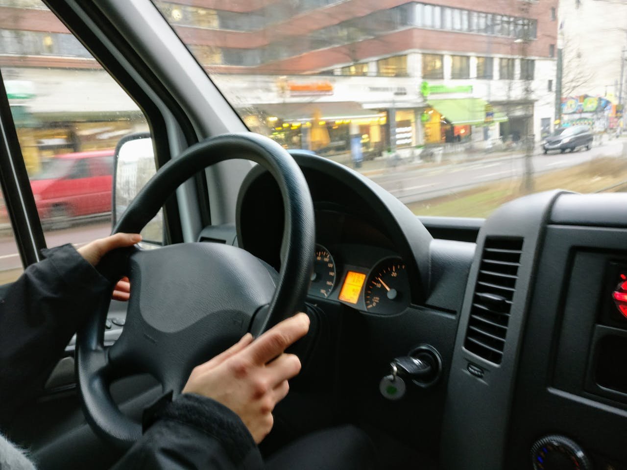 Servicios Hands on a steering wheel driving a van through an urban area with shops visible outside.