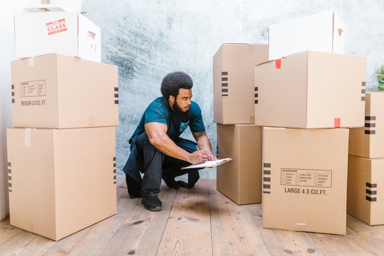 Servicios An adult man in workwear checks a clipboard while surrounded by moving boxes in an indoor setting.
